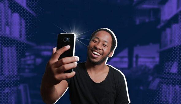 A man wearing a black t-shirt against a dark purple background. He is smiling at his phone while taking a selfie.