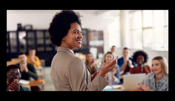 A female educator with dark skin, dark hair, and a tan blazer smiling as she teaches her students, indicating her overall well-being.