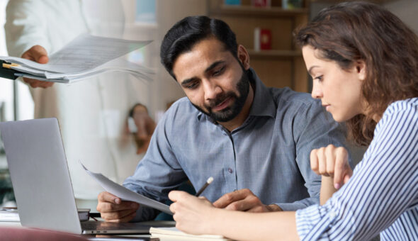 Two people collaborating at a desk in an office setting. A bearded man in a gray shirt reviews a document with a young woman in a striped shirt who appears focused on taking notes. A laptop and notebook are on the desk in front of them.