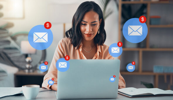 What speaks to your donors? Emojis, GIFS, smoke signals? article image. A woman is seated at a desk, working on a laptop in an office setting. She is surrounded by floating icons of envelopes with red dollar signs, symbolizing email fundraising or alumni giving donations.