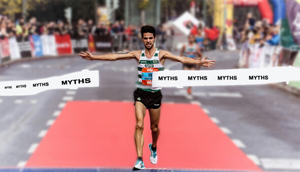 A male runner with short dark hair and beard, wearing a white and black striped shirt and black shorts running through a race banner with the word "Myth" written across it multiple times.