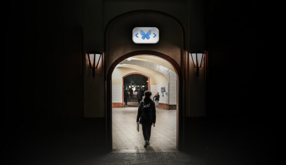 A male college student walking through an open doorway with an "exit" sign above. Instead of "exit" the sign depicts the blue butterfly of the Bluesky logo.