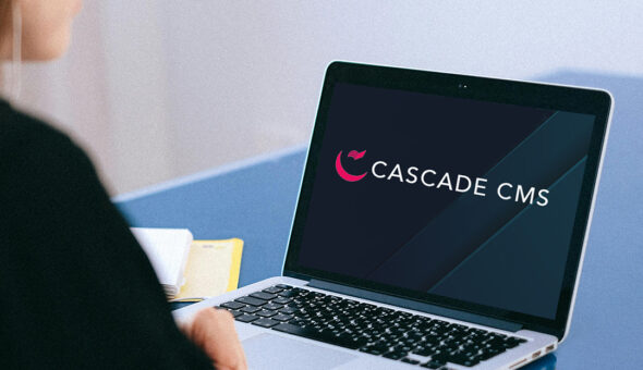 What’s next for Cascade CMS? article image. A person sits at a desk using a laptop displaying the Cascade CMS logo on the screen. Nearby, there are a few papers and a yellow folder. The setting appears to be an office or workspace.