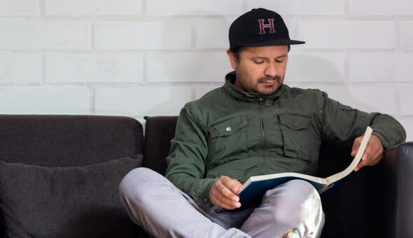 Do alumni magazines still matter? article image. Man sitting on a dark couch reading a book, wearing a green jacket and a black baseball cap with a red letter "H" on it, against a white brick wall background.