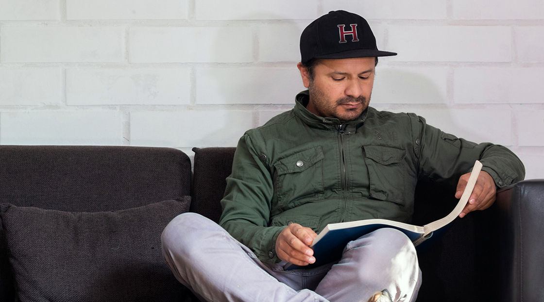image - Do alumni magazines still matter? article image. Man sitting on a dark couch reading a book, wearing a green jacket and a black baseball cap with a red letter "H" on it, against a white brick wall background.