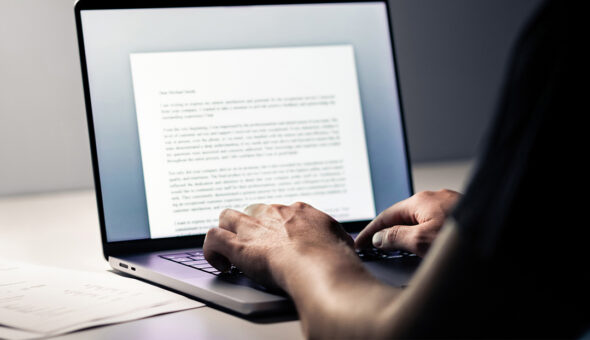 PR Writing Tips for Great Pitches article title. Person typing on a laptop with a document open on the screen, showing a partially visible letter. The setting appears to be a dimly lit workspace with papers on the desk.