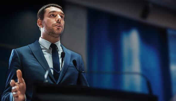 Leadership Communications Tips for Higher Ed article image. A man in a suit speaking confidently at a podium with two microphones, in front of a blurred blue backdrop at what appears to be a conference or formal event.