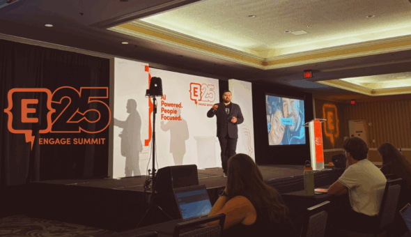 A speaker stands on stage at the Engage Summit 2025 in a hotel conference room, presenting in front of a branded backdrop that reads "AI Powered. People Focused." and "E25 Engage Summit." Attendees are seated in the audience, engaged and taking notes on laptops.